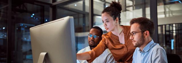 Mulher branca, entre um homem branco e um homem negro, apontando para um monitor de computador.